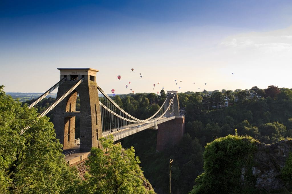 A suspension bridge spans over a gorge, surrounded by lush greenery. Numerous hot air balloons float in the sky during a sunny day, creating a picturesque scene, while nearby residential scaffolding highlights ongoing construction amidst nature's splendor.