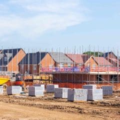 A residential construction site with several new brick houses, scaffolding, construction materials, and yellow machinery under a clear blue sky.