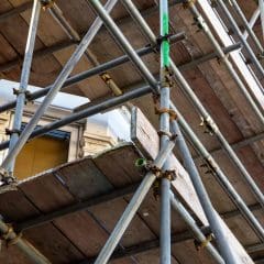 Metal scaffolding is set up alongside a brick building, with wooden planks forming walkways at different levels. A window partially covered by the structure is visible in the background.