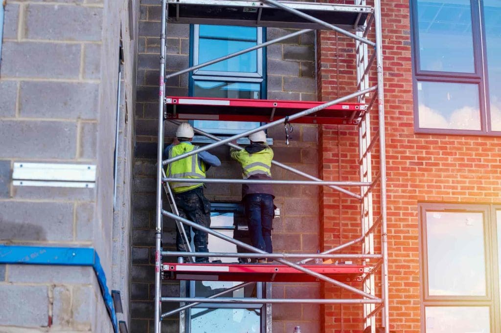 Two construction workers in safety vests and helmets are standing on scaffolding, working on the exterior wall of a building with brick and concrete blocks, near large windows.